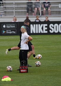 A person in sports attire stands on a soccer field, surrounded by several soccer balls and a carrier with bottles. The field is bordered by advertisements for Nike Soccer and app stores. Several people sit in bleachers in the background.