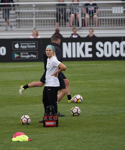 A person in sports attire stands on a soccer field, surrounded by several soccer balls and a carrier with bottles. The field is bordered by advertisements for Nike Soccer and app stores. Several people sit in bleachers in the background.