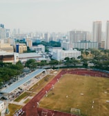An aerial view of a sports complex with a running track surrounding a grassy field, set against the background of a cityscape. The city's skyline features a mix of tall residential buildings and shorter commercial structures. There are trees and green areas surrounding the sports facility.