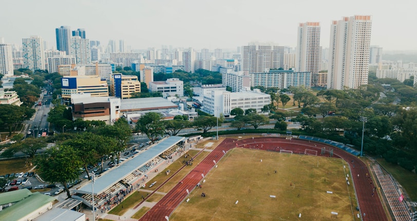 An aerial view of a sports complex with a running track surrounding a grassy field, set against the background of a cityscape. The city's skyline features a mix of tall residential buildings and shorter commercial structures. There are trees and green areas surrounding the sports facility.