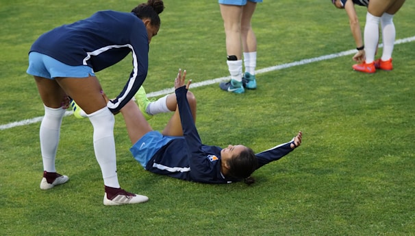 A group of athletes in sportswear are on a green soccer field. Two individuals are interacting closely; one helps the other who is lying on the grass. Nearby, other athletes are stretching.