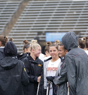 A group of people, likely athletes, are gathered together on a sports field. They are wearing black jackets with some individuals dressed in white sports uniforms underneath. A few people have their hair tied up in buns, and most are engaged in conversation. The background shows tiered stadium seats, suggesting the setting is an outdoor sports venue.