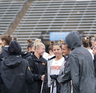 A group of people, likely athletes, are gathered together on a sports field. They are wearing black jackets with some individuals dressed in white sports uniforms underneath. A few people have their hair tied up in buns, and most are engaged in conversation. The background shows tiered stadium seats, suggesting the setting is an outdoor sports venue.