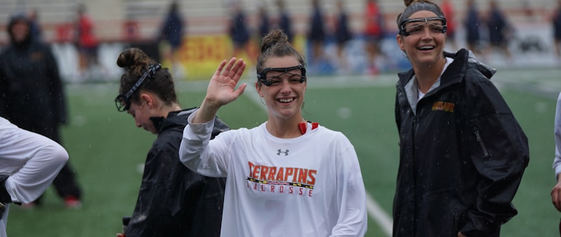 Several athletes in sports attire are on an outdoor sports field, interacting and smiling. One individual in the foreground waves with a friendly expression, while another stands beside her, also smiling. They wear protective goggles and gear associated with lacrosse. The background features indistinct figures in dark clothing against a backdrop of stadium seating.