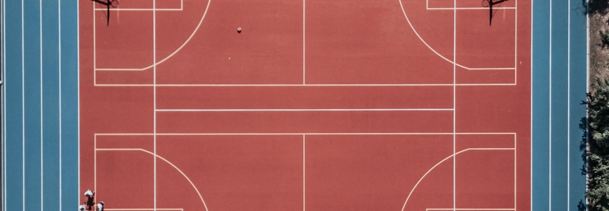 An aerial view of a large, vibrant sports complex featuring a red basketball court with multiple hoops and a surrounding blue running track. Several people are scattered across the area, engaged in various activities.