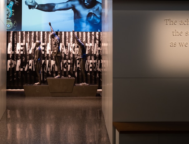 A dimly lit display features statues of athletes standing on a podium. They are positioned in front of a backdrop with the words 'LEVELING THE PLAYING FIELD' in bold letters. On the wall to the right, a softly lit quote is partially visible. The overall ambiance suggests a museum or exhibition setting.