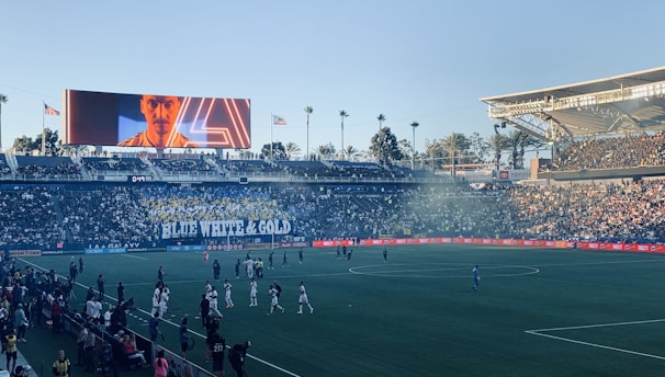 A large sports stadium filled with spectators. The field is occupied by soccer players, some walking and others scattered across the pitch. A massive screen displays a person, and flags fly on top of the stadium roof. The audience is dense, and the atmosphere seems lively with a visible banner showing 'BLUE WHITE & GOLD'.
