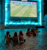 A large outdoor screen displays a soccer match at night. A small group of people sit on a cobblestone surface, watching the game. The screen is surrounded by a bright blue glow, creating a lively atmosphere.