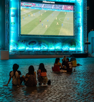 A large outdoor screen displays a soccer match at night. A small group of people sit on a cobblestone surface, watching the game. The screen is surrounded by a bright blue glow, creating a lively atmosphere.