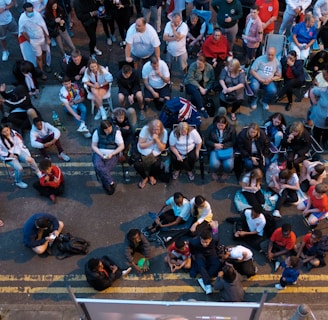 A large group of people gathered outdoors, watching a screen that is displaying a man. The crowd is diverse in age and appears interested and engaged, with many sitting closely together, some standing. Many are dressed casually, some in sports attire, and a few waving or holding flags. The event seems to have a communal, festive atmosphere.