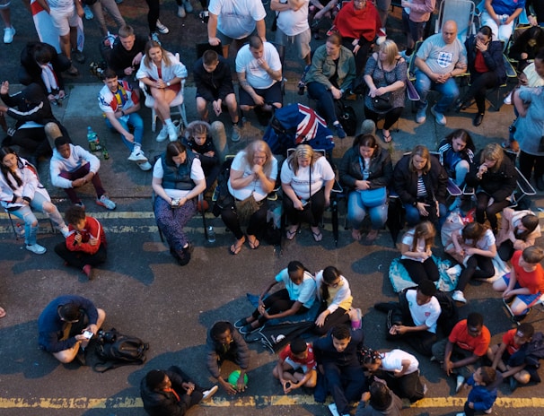 A large group of people gathered outdoors, watching a screen that is displaying a man. The crowd is diverse in age and appears interested and engaged, with many sitting closely together, some standing. Many are dressed casually, some in sports attire, and a few waving or holding flags. The event seems to have a communal, festive atmosphere.
