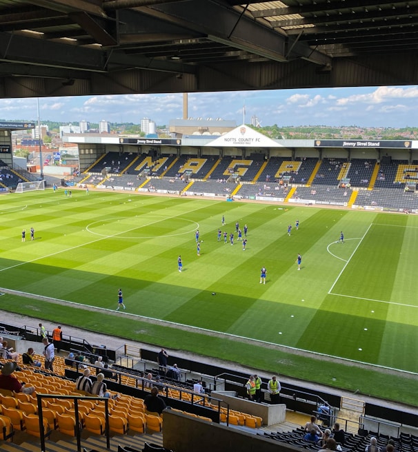 A sports stadium with a well-maintained grass field is depicted. Players are spread across the field, engaging in warm-up activities. The stands are partially filled with spectators, and the stadium is surrounded by urban buildings under a partly cloudy sky.