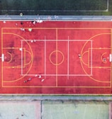An aerial view of a sports court with a vibrant red surface and yellow lines marking different sections for basketball and soccer. Several people are present on the court, some engaged in activities, while others are standing along the perimeter. The court is enclosed by a metal fence and surrounded by grass on all sides.