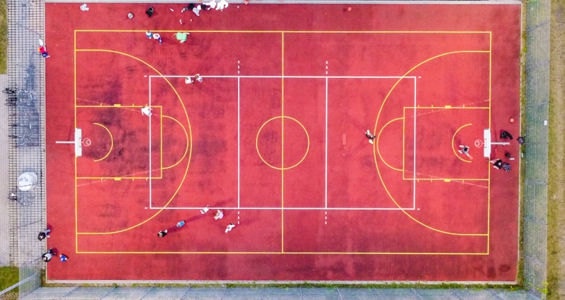 An aerial view of a sports court with a vibrant red surface and yellow lines marking different sections for basketball and soccer. Several people are present on the court, some engaged in activities, while others are standing along the perimeter. The court is enclosed by a metal fence and surrounded by grass on all sides.