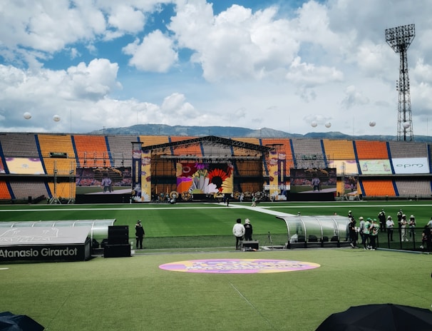 A sports stadium with a large stage set up on the field, hosting an outdoor event. Spectators wearing colorful hats sit in the foreground. The seating areas display vibrant colors, and a clear blue sky with scattered clouds is visible. Large screens display graphics related to the event.