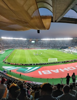 A large, brightly lit stadium filled with spectators. The field is green and well-maintained, with players actively engaged in a soccer match. The stands are crowded, and banners with writing are visible along the perimeter of the field. The atmosphere suggests excitement and energy.