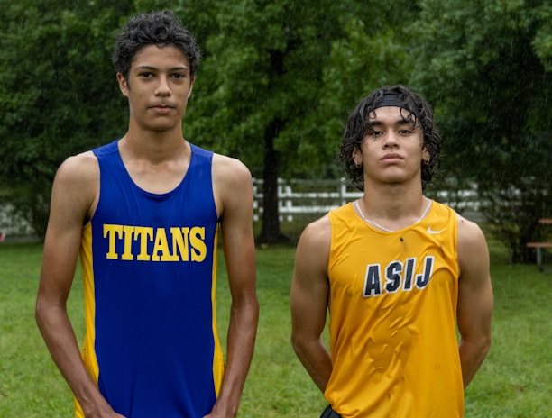 Two young athletes wearing sleeveless sports tops stand on a grassy field. The athlete on the left is wearing a blue top with the word 'TITANS' while the one on the right wears a yellow top with 'ASIJ'. The background features green trees and a white fence, with a bench visible on the right.