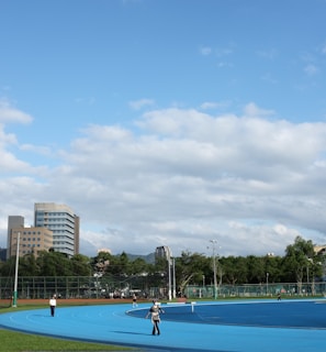 A bright outdoor sports complex features a blue running track, surrounded by green grass and trees. Several people are visible, engaged in activities on the track. In the background, urban high-rise buildings rise against a partly cloudy sky.