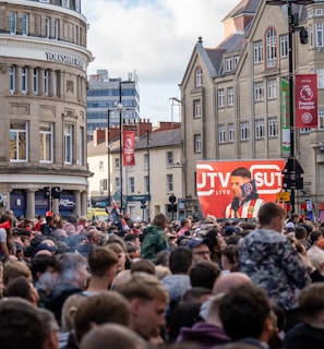 A large crowd of people, many wearing red and white, gather in a city square. They are watching an event broadcast on a large screen, which displays a man speaking into a microphone with logos suggesting a sporting event. Surrounding buildings are historical and commercial, with banners indicating a connection to the Premier League.
