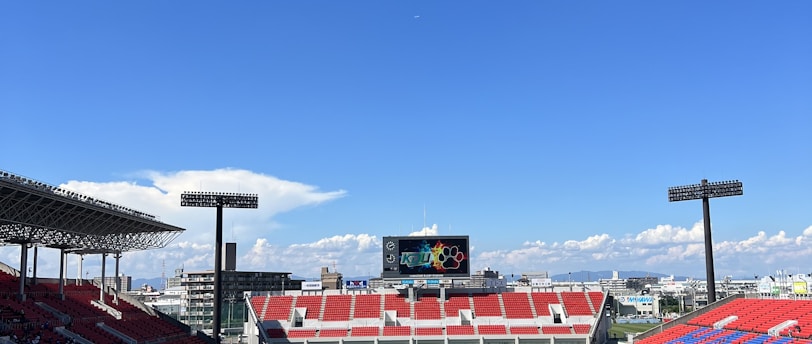 A large sports stadium with red and blue seating overlooks a green field with a goalpost at the center. A few people are scattered across the field, with some tents and a gathering of individuals on the right side. The sky is clear and blue with some clouds visible in the distance. A large screen or scoreboard is situated at the far end of the field.