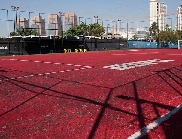 A sports court with a red playing surface and white lines, surrounded by a black mesh fence. In the background, tall buildings are visible, along with some trees and a few yellow chairs positioned near the fence.
