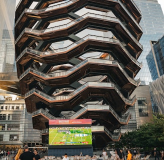 A large, spiral architectural structure with multiple levels stands prominently in an urban setting. In front of the structure, a large screen displays a sports broadcast, and several people are gathered around, some sitting and others standing. The scene is set outdoors, with towering modern buildings surrounding the area. The ground is paved with a geometric pattern of gray and white tiles.