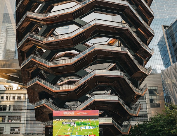 A large, spiral architectural structure with multiple levels stands prominently in an urban setting. In front of the structure, a large screen displays a sports broadcast, and several people are gathered around, some sitting and others standing. The scene is set outdoors, with towering modern buildings surrounding the area. The ground is paved with a geometric pattern of gray and white tiles.